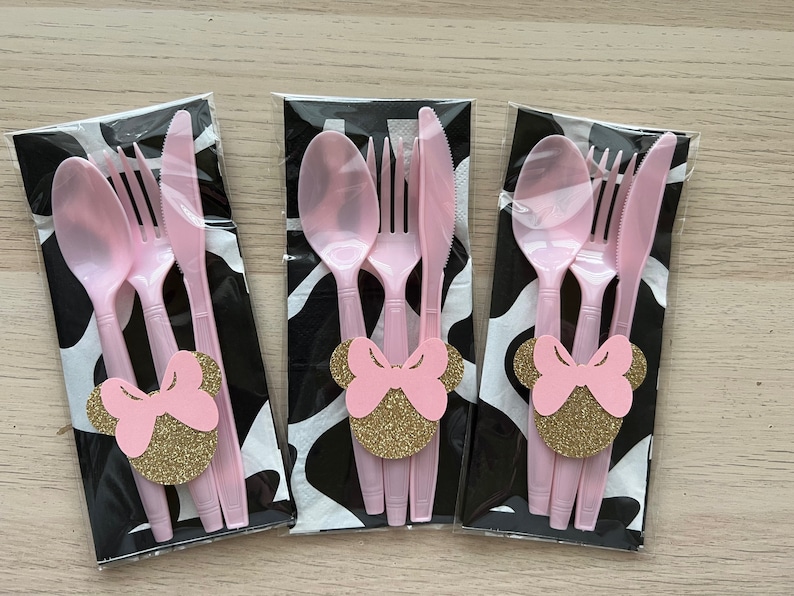 Puede incluir: Tres juegos de cubiertos de pl&aacute;stico rosa, cada juego incluye un tenedor, un cuchillo y una cuchara. Cada juego est&aacute; envuelto en una servilleta negra y blanca con un recorte de cabeza de Minnie Mouse rosa y dorado.