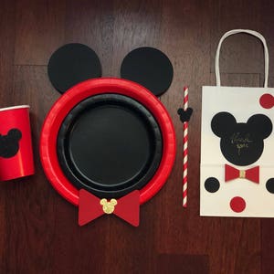 Puede incluir: Un conjunto de art&iacute;culos para fiestas con tem&aacute;tica de Mickey Mouse. Incluye una taza roja, platos y una bolsa de regalo con pajarita y la frase "gracias". El conjunto tambi&eacute;n tiene una pajita y orejas de rat&oacute;n negras.