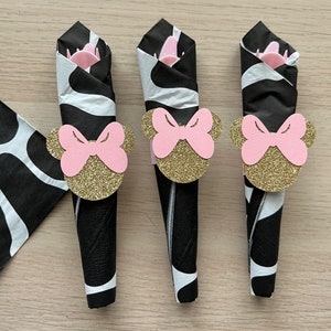 Puede incluir: Tres servilletas negras y blancas con anillos de servilleta de cabeza de Minnie Mouse con purpurina dorada. Los anillos de servilleta tienen lazos rosas.