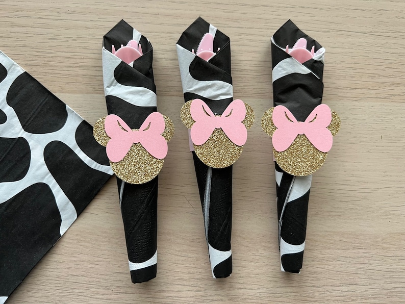 Puede incluir: Tres servilletas negras y blancas con anillos de servilleta de cabeza de Minnie Mouse con purpurina dorada. Los anillos de servilleta tienen lazos rosas.