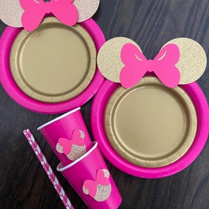 Puede incluir: Dos platos de papel rosa con orejas de Mickey Mouse de purpurina dorada y lazos rosas. Dos vasos de papel rosa con orejas de Mickey Mouse de purpurina dorada y lazos rosas. Dos pajitas de papel a rayas.