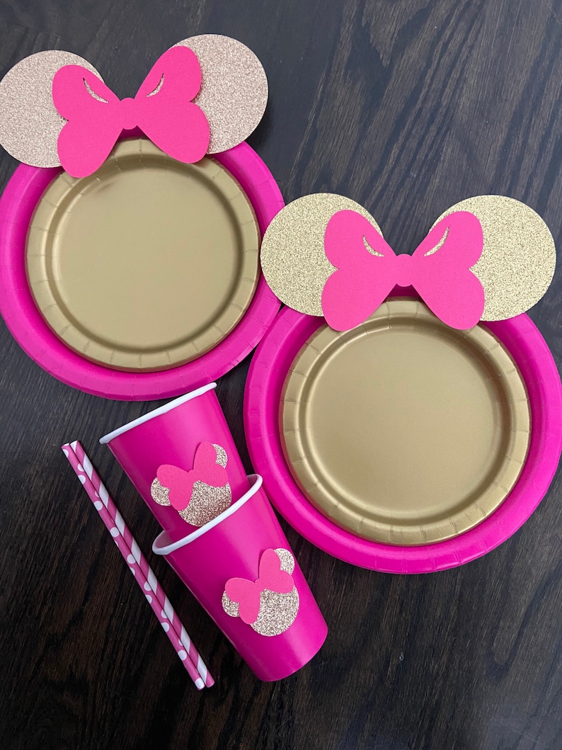 Puede incluir: Dos platos de papel rosa con orejas de Mickey Mouse de purpurina dorada y lazos rosas. Dos vasos de papel rosa con orejas de Mickey Mouse de purpurina dorada y lazos rosas. Dos pajitas de papel a rayas.