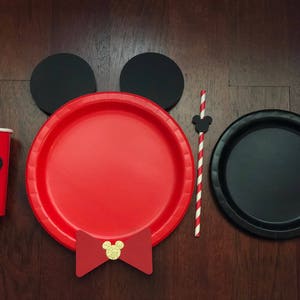 Puede incluir: Un juego de fiesta festivo con una taza y un plato rojos con una silueta negra de Mickey Mouse, recortes de orejas negras, una pajarita roja y un plato negro. Tambi&eacute;n se incluye una pajita a rayas rojas y blancas con un recorte de Mickey Mouse.
