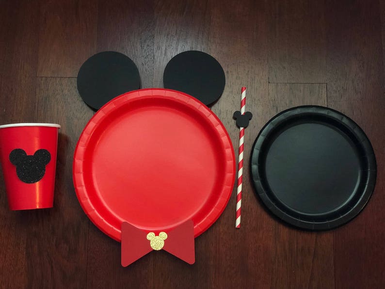 Puede incluir: Un juego de fiesta festivo con una taza y un plato rojos con una silueta negra de Mickey Mouse, recortes de orejas negras, una pajarita roja y un plato negro. Tambi&eacute;n se incluye una pajita a rayas rojas y blancas con un recorte de Mickey Mouse.