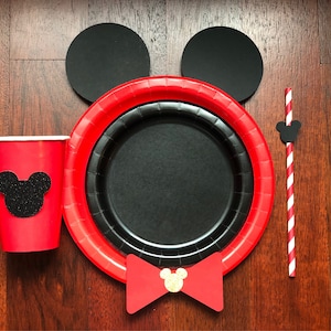 Puede incluir: Un juego de fiesta festivo con una taza roja con una silueta de rat&oacute;n de purpurina negra, un plato rojo y negro, un plato negro, dos c&iacute;rculos negros, una pajita a rayas rojas y blancas y una pajarita roja con una cabeza de rat&oacute;n dorada.