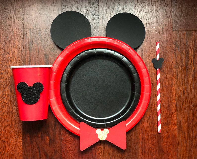 Puede incluir: Un juego de fiesta festivo con una taza roja con una silueta de rat&oacute;n de purpurina negra, un plato rojo y negro, un plato negro, dos c&iacute;rculos negros, una pajita a rayas rojas y blancas y una pajarita roja con una cabeza de rat&oacute;n dorada.