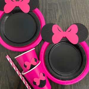 Puede incluir: Platos y vasos de papel rosa y negro con un dise&ntilde;o de Minnie Mouse. Los platos tienen un centro negro y un borde rosa. Los vasos tienen una base rosa y una cabeza de Minnie Mouse negra con un lazo rosa en la parte superior.