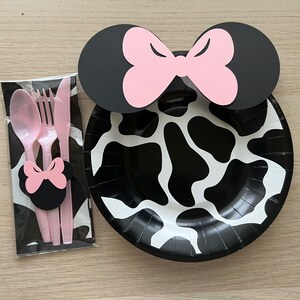 Puede incluir: Un plato de papel de vaca en blanco y negro con un lazo de Minnie Mouse rosa y negro en la parte superior. El plato es parte de un juego de suministros para fiestas que incluye una cuchara, un tenedor y un cuchillo de pl&aacute;stico rosa.