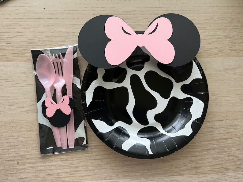 Puede incluir: Un plato de papel de vaca en blanco y negro con un lazo de Minnie Mouse rosa y negro en la parte superior. El plato es parte de un juego de suministros para fiestas que incluye una cuchara, un tenedor y un cuchillo de pl&aacute;stico rosa.