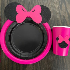 Puede incluir: Suministros para fiestas de Minnie Mouse en rosa y negro. Un plato de papel rosa con un plato de papel negro en el interior, una taza rosa con una cabeza de Minnie Mouse negra y un lazo, y una pajita de lunares rosa y blanca.
