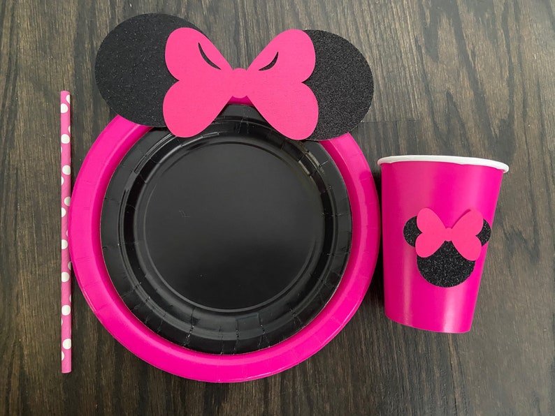 Puede incluir: Suministros para fiestas de Minnie Mouse en rosa y negro. Un plato de papel rosa con un plato de papel negro en el interior, una taza rosa con una cabeza de Minnie Mouse negra y un lazo, y una pajita de lunares rosa y blanca.