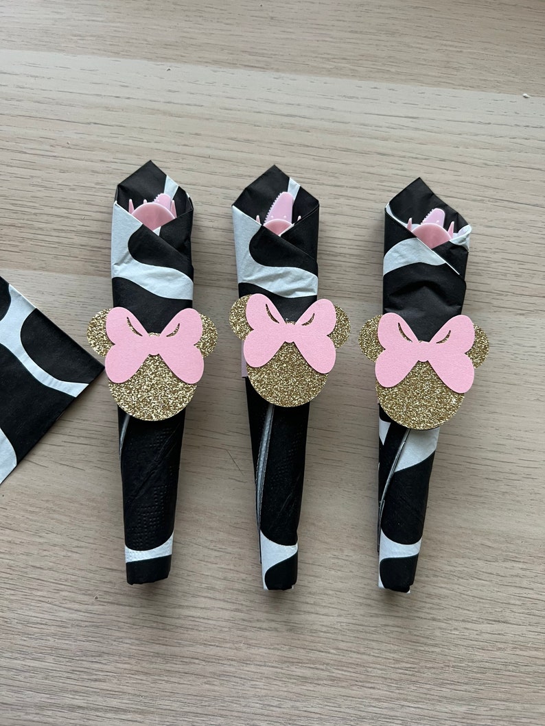 Puede incluir: Tres servilletas de papel con estampado de cebra en blanco y negro con anillos de servilleta de lazo rosa y dorado. Los anillos de lazo tienen un acabado brillante.