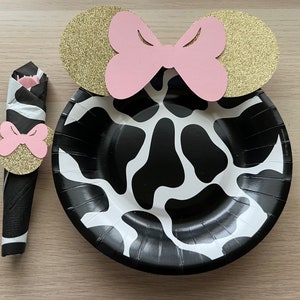 Puede incluir: Un plato de papel de vaca en blanco y negro con un anillo de servilleta de orejas de Minnie Mouse doradas y rosadas. El anillo de servilleta tiene un lazo rosa y purpurina dorada.