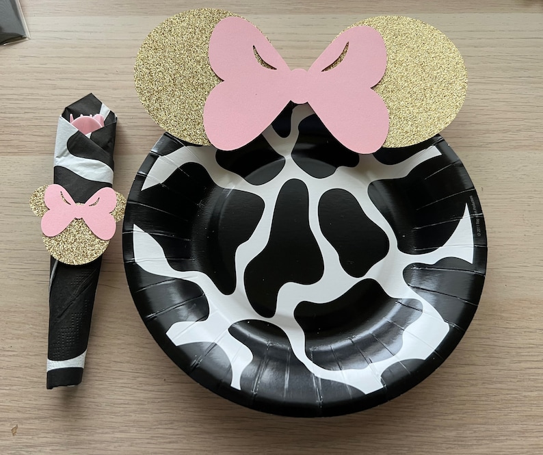 Puede incluir: Un plato de papel de vaca en blanco y negro con un anillo de servilleta de orejas de Minnie Mouse doradas y rosadas. El anillo de servilleta tiene un lazo rosa y purpurina dorada.