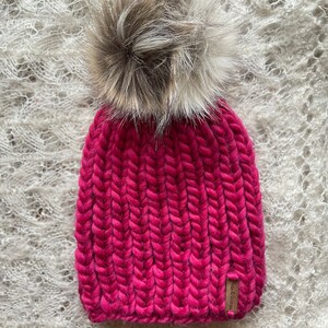 May include: A pink knitted beanie with a fluffy brown pom pom. The beanie has a brown leather tag with the text "Wool & Grace" on it.