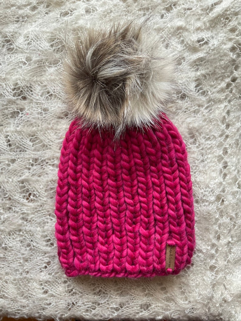 May include: A pink knitted beanie with a fluffy brown pom pom. The beanie has a brown leather tag with the text "Wool & Grace" on it.
