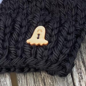 Op de afbeelding: Een zwarte gebreide muts met een houten spookknop. De knop is lichtbruin met een wit gezicht en zwarte ogen.