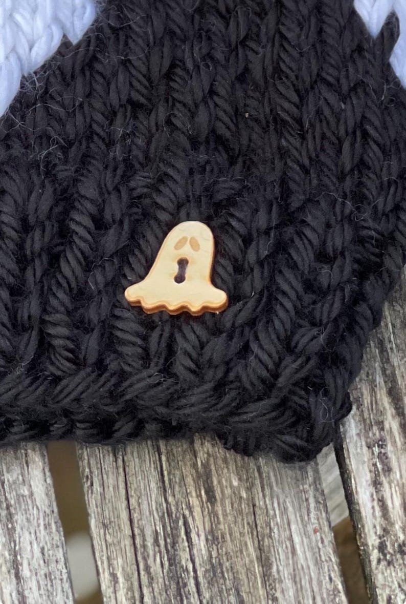 Op de afbeelding: Een zwarte gebreide muts met een houten spookknop. De knop is lichtbruin met een wit gezicht en zwarte ogen.