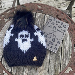 Op de afbeelding: Zwart-witte gebreide muts met een spookontwerp en een zwarte pompon. De muts heeft een houten knoop in de vorm van een spook. De muts ligt op een houten tafel met een notitieboekje waarop "Hugs & Stitches" staat.