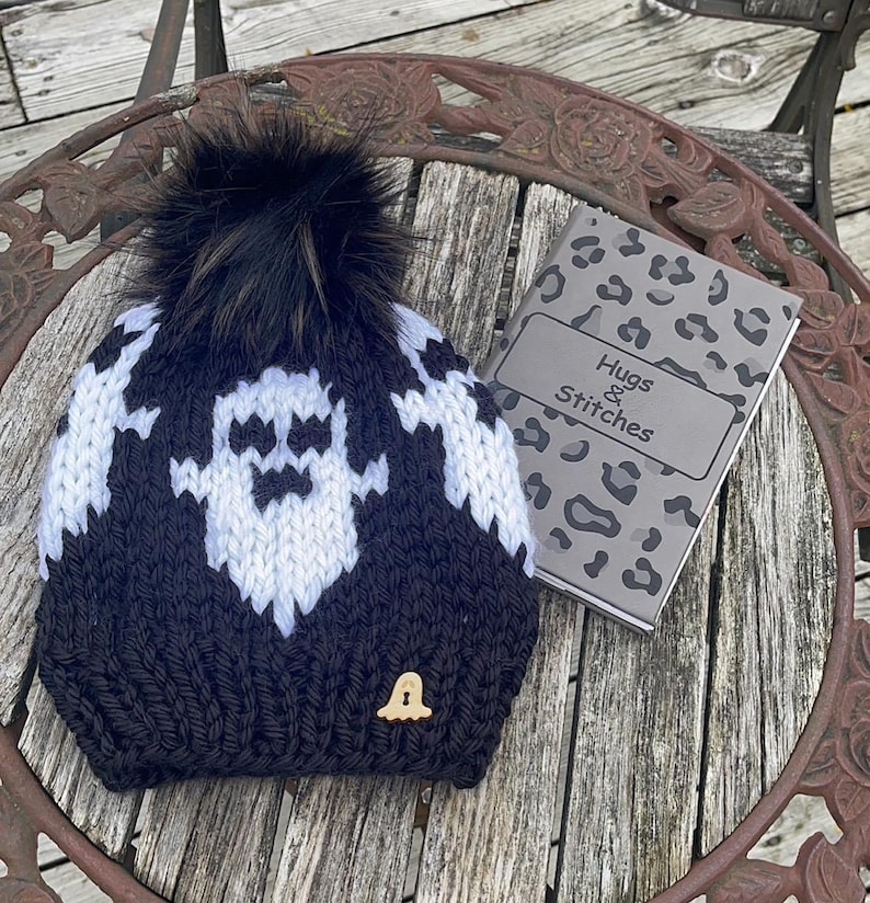Op de afbeelding: Zwart-witte gebreide muts met een spookontwerp en een zwarte pompon. De muts heeft een houten knoop in de vorm van een spook. De muts ligt op een houten tafel met een notitieboekje waarop "Hugs & Stitches" staat.