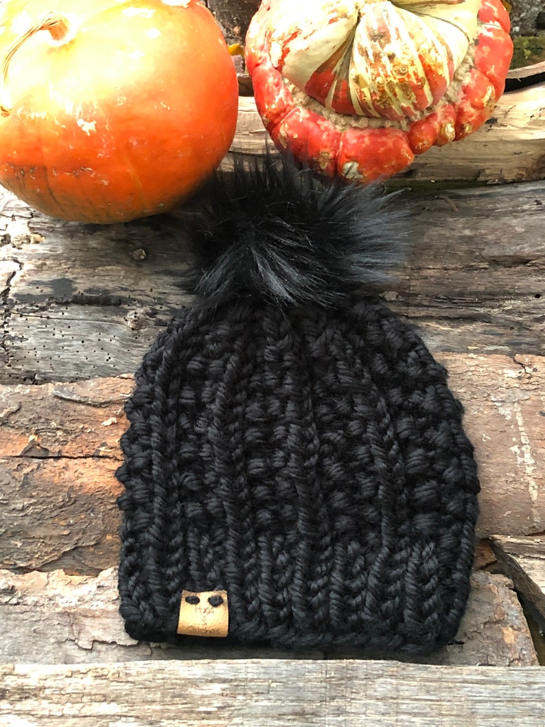 May include: Black knitted beanie with a fluffy pom pom and a brown leather tag that says "Hope & Stitches".