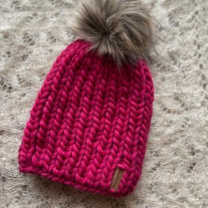 May include: A pink knitted beanie with a faux fur pom pom. The beanie has a brown leather tag with the text "Wool & Grace" on it.