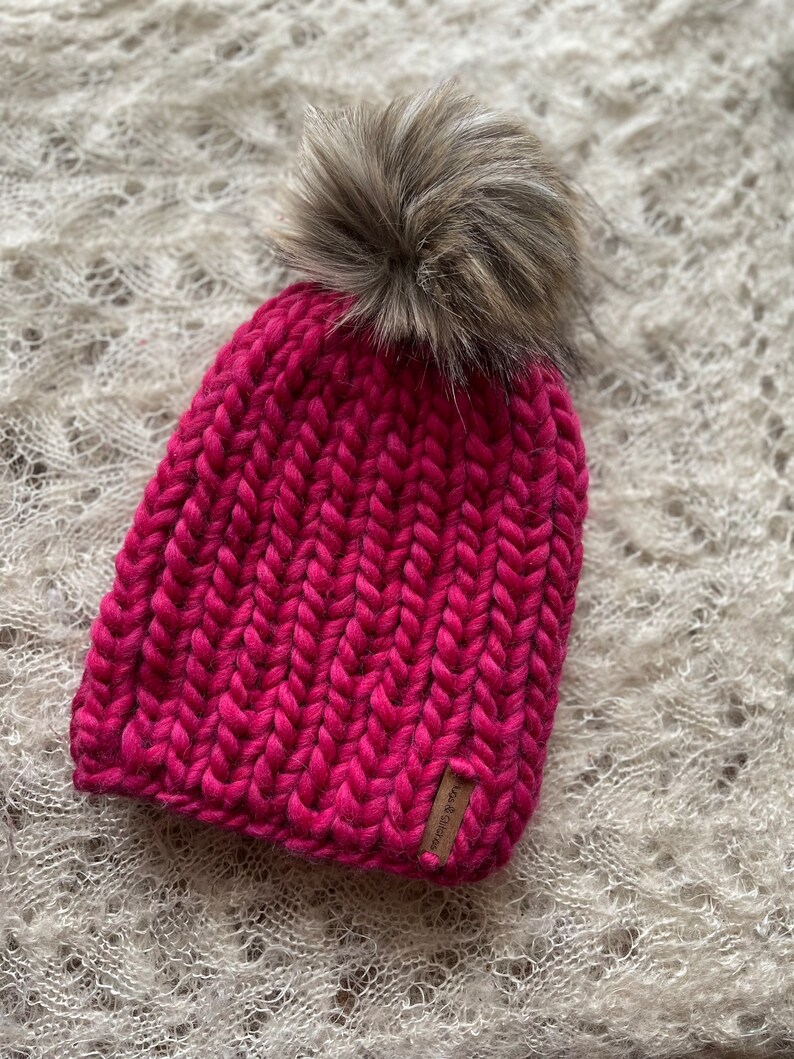 May include: A pink knitted beanie with a faux fur pom pom. The beanie has a brown leather tag with the text "Wool & Grace" on it.
