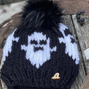 Op de afbeelding: Zwart-witte gebreide beanie met een wit spookontwerp en een zwarte nep bont pompon. De beanie heeft een houten spookknop.