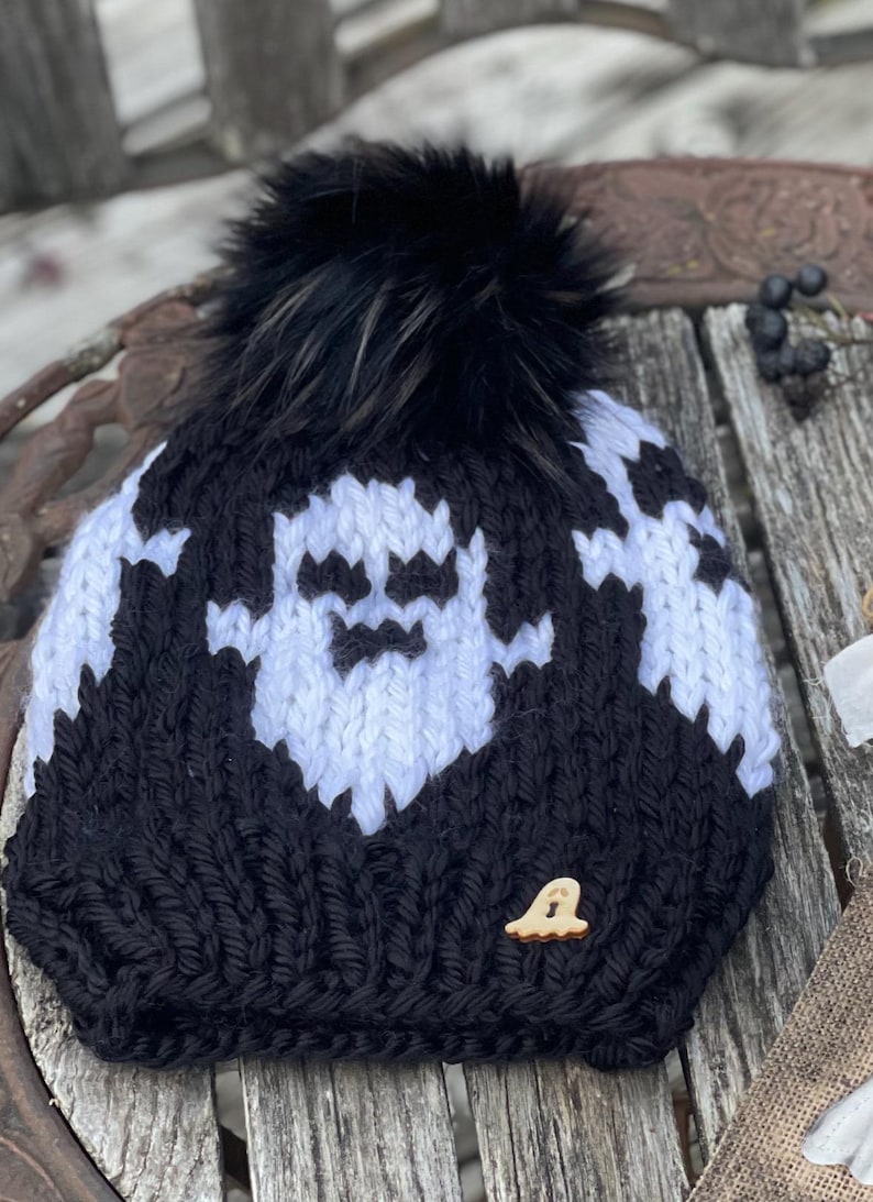 Op de afbeelding: Zwart-witte gebreide beanie met een wit spookontwerp en een zwarte nep bont pompon. De beanie heeft een houten spookknop.