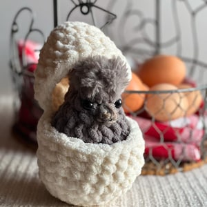 Puede incluir: Un huevo blanco de crochet con un pollito gris esponjoso asomando por la parte superior. El huevo está sobre una superficie blanca.