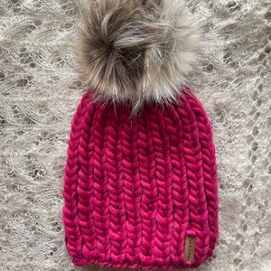 May include: A pink knitted beanie with a fluffy brown pom pom. The beanie has a ribbed texture and a small brown leather tag with the word "Handmade" on it.