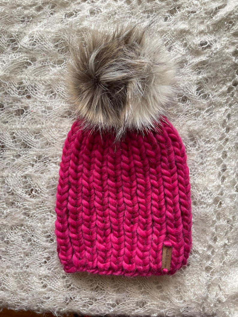 May include: A pink knitted beanie with a fluffy brown pom pom. The beanie has a ribbed texture and a small brown leather tag with the word "Handmade" on it.
