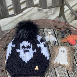 Op de afbeelding: Zwart-witte gebreide beanie met een zwarte nep bont pompon en een spookontwerp. De beanie heeft een kleine houten knoop met een spookontwerp aan de zijkant. De beanie ligt op een houten tafel met een jute spookbanner.