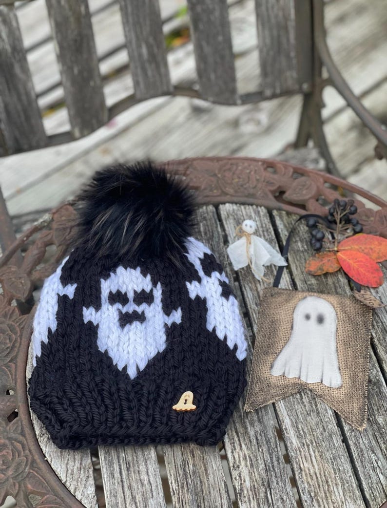 Op de afbeelding: Zwart-witte gebreide beanie met een zwarte nep bont pompon en een spookontwerp. De beanie heeft een kleine houten knoop met een spookontwerp aan de zijkant. De beanie ligt op een houten tafel met een jute spookbanner.