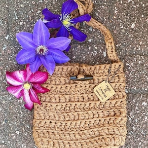 May include: A brown crocheted bag with a long strap. The bag has a wooden button closure and a small wooden tag that says "Hugs & Stitches Handmade in WI". The bag is surrounded by purple and pink flowers.