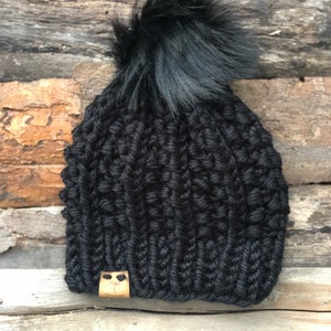May include: Black knitted beanie with a fluffy black pom pom and a brown leather tag that says "Hugs & Stitches".
