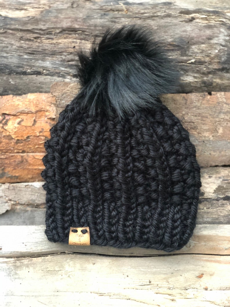 May include: Black knitted beanie with a fluffy black pom pom and a brown leather tag that says "Hugs & Stitches".