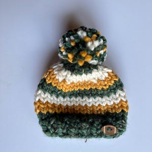 Puede incluir: Un gorro de punto a rayas verde, blanco y amarillo con un pompón en la parte superior y un botón de madera en el lateral.