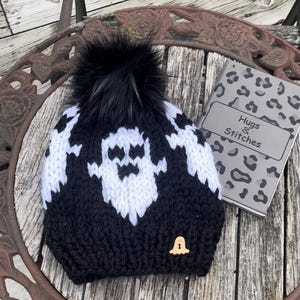 Op de afbeelding: Zwart-witte gebreide beanie met een spookontwerp en een zwarte nep bont pompon. De beanie heeft een klein houten belletje eraan bevestigd. De beanie ligt op een houten tafel met een notitieboekje waarop "Hugs & Stitches" staat.