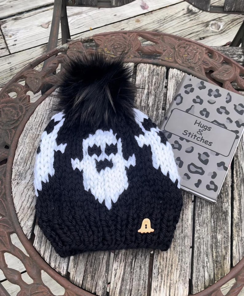 Op de afbeelding: Zwart-witte gebreide beanie met een spookontwerp en een zwarte nep bont pompon. De beanie heeft een klein houten belletje eraan bevestigd. De beanie ligt op een houten tafel met een notitieboekje waarop "Hugs & Stitches" staat.