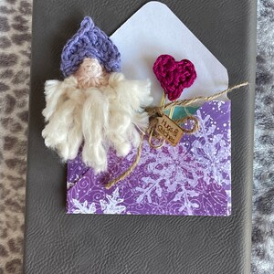 May include: A purple crocheted gnome with a white beard and a pink heart-shaped paper clip attached to a purple envelope with a white snowflake pattern. The envelope has a brown tag that says "Hugs & Stitches".