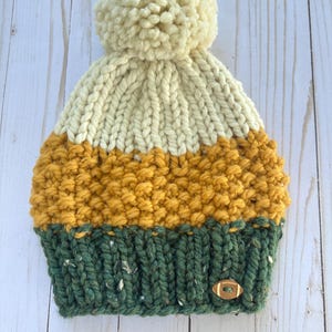 May include: A hand-knitted beanie in cream, mustard yellow, and forest green. The hat features a large cream-colored pom-pom on top and a small wooden football-shaped button on the brim. The hat is made of thick, textured yarn.