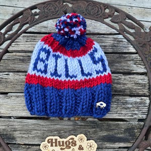 Può includere: Un berretto lavorato a maglia blu, rosso e bianco con un pompon. Il berretto presenta la scritta "BILLS" in bianco, una striscia rossa e un piccolo emblema di bufalo. Il cartello di legno sottostante recita "Hugs & Stitches".