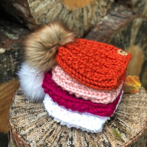 May include: A stack of four knitted hats with pom-poms. The hats are in shades of orange, pink, red, and white.
