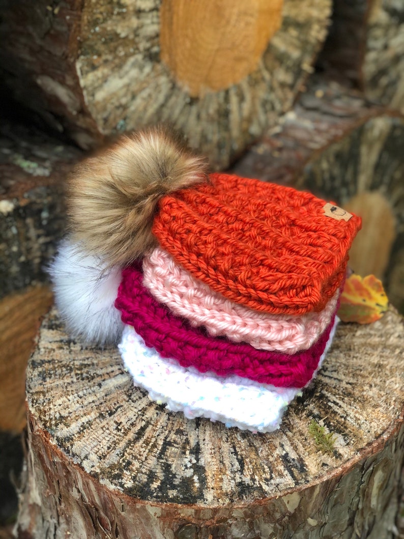 May include: A stack of four knitted hats with pom-poms. The hats are in shades of orange, pink, red, and white.