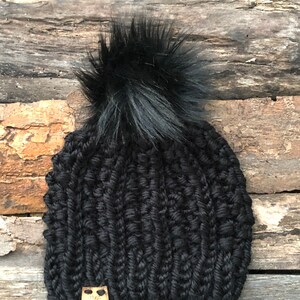 May include: Black knitted beanie with a large black pom pom and a small brown leather tag with a logo.