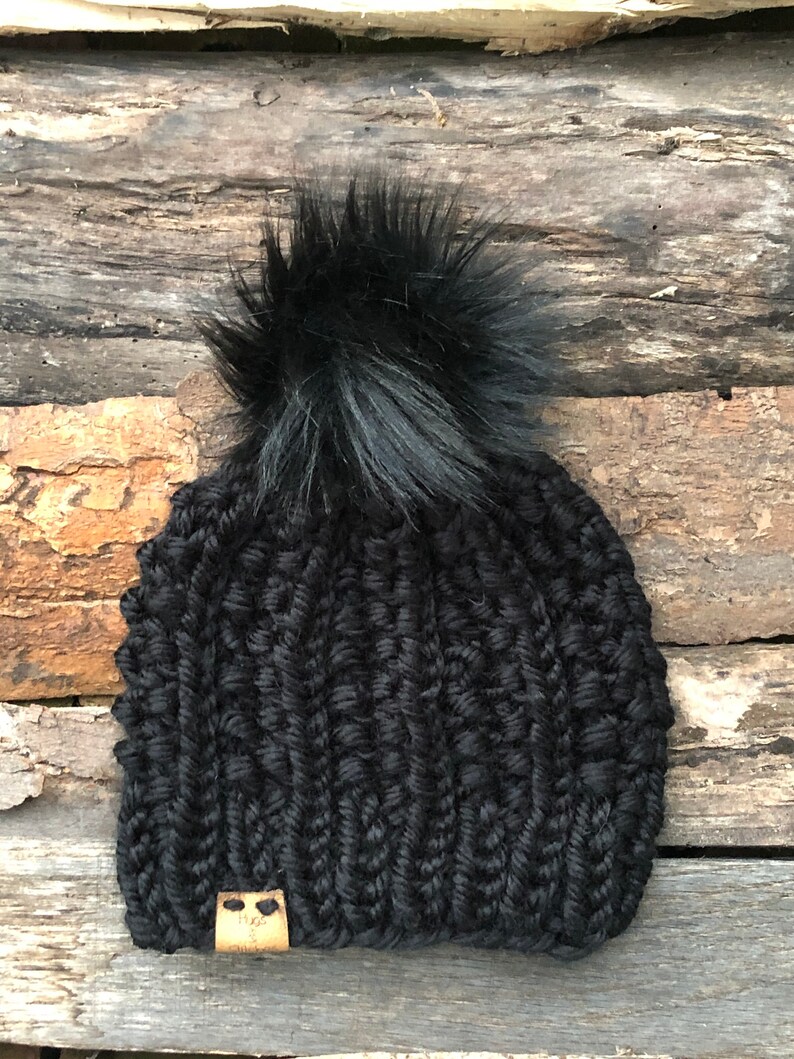 May include: Black knitted beanie with a large black pom pom and a small brown leather tag with a logo.