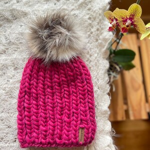 May include: A pink knitted beanie with a fluffy brown pom pom. The beanie has a brown leather tag with the text "Wool & Glaze" on it.