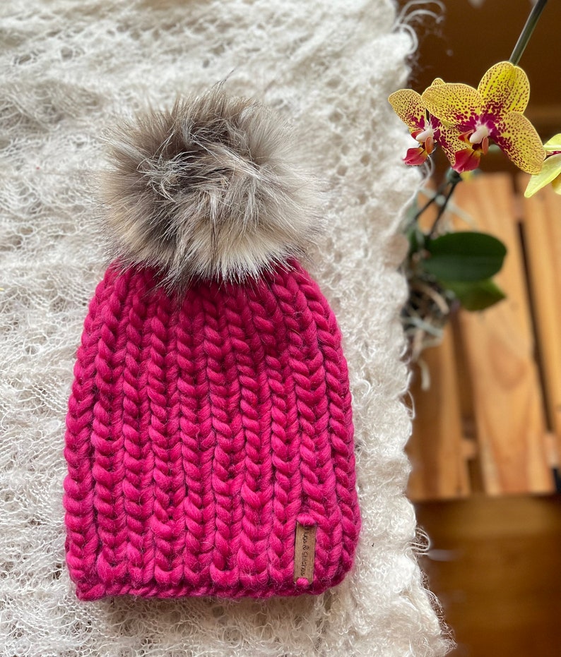 May include: A pink knitted beanie with a fluffy brown pom pom. The beanie has a brown leather tag with the text "Wool & Glaze" on it.