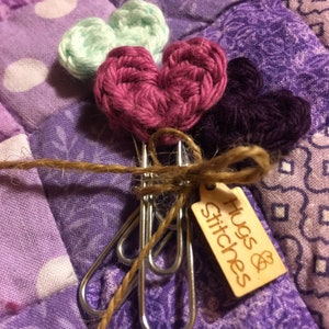 Puede incluir: Tres corazones de ganchillo en tonos de morado, verde y rosa están unidos a un juego de clips de carpeta plateados. Los clips están atados con un cordel y una etiqueta de madera que dice "Hugs & Stitches".